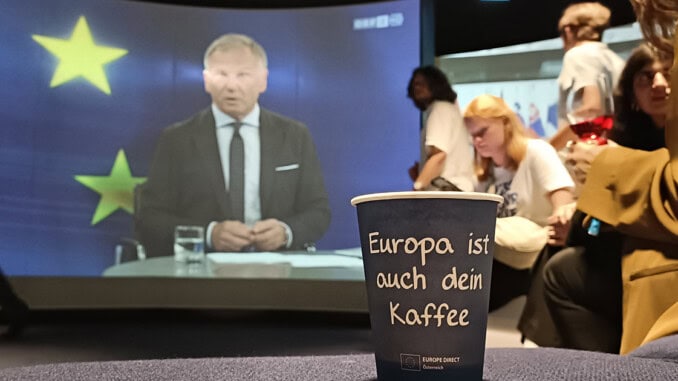 Kaffeebecher mit der Aufschrift: „Europa ist auch dein Kafee“ und Armin Wolf auf einer Kinoleinwand im Hintergrund